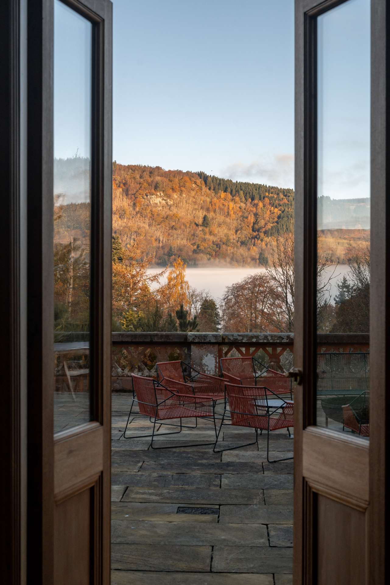 Open French doors frame a view of red wicker chairs on a stone terrace overlooking misty autumn hills and a loch.