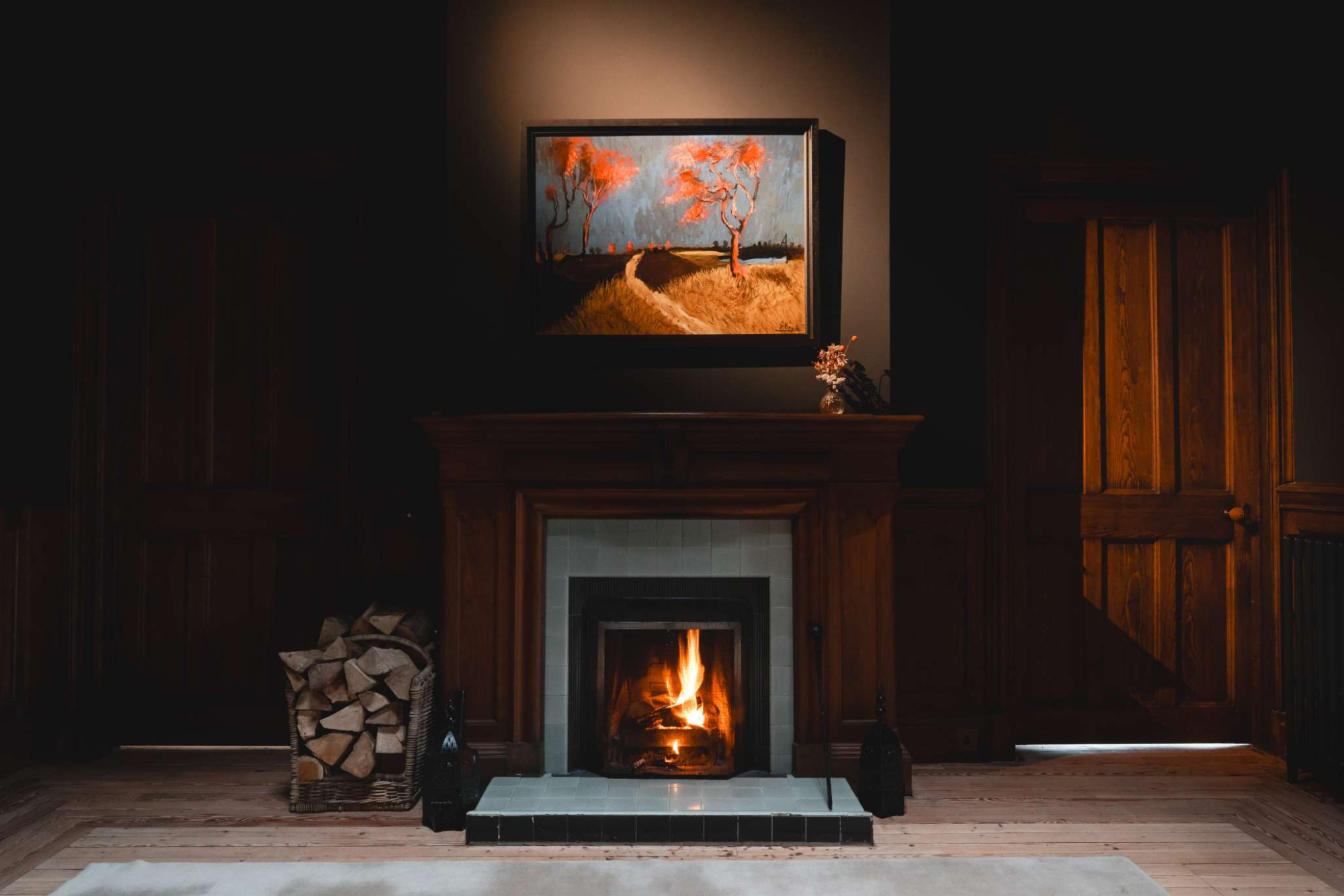 A wood-panelled fireplace with a lit fire sits beneath a framed painting of red-blossomed trees, an open doorway visible to the side.