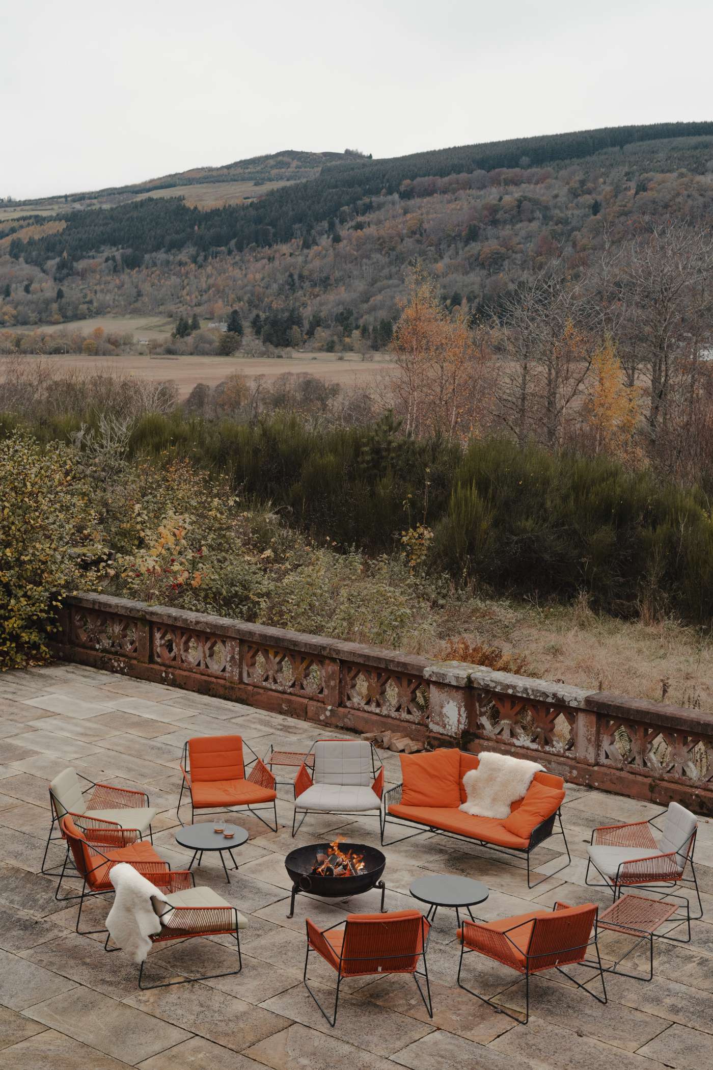 Orange and grey chairs are arranged around a lit firepit on a stone terrace with a carved balustrade, autumn trees and hills beyond.