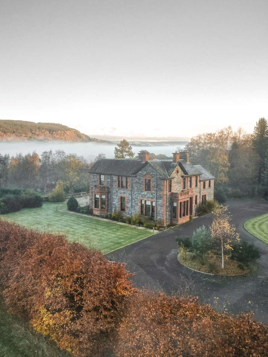 An aerial view shows a stone and sandstone house set in a lawned garden with a curved driveway, mist filling the valley beyond.