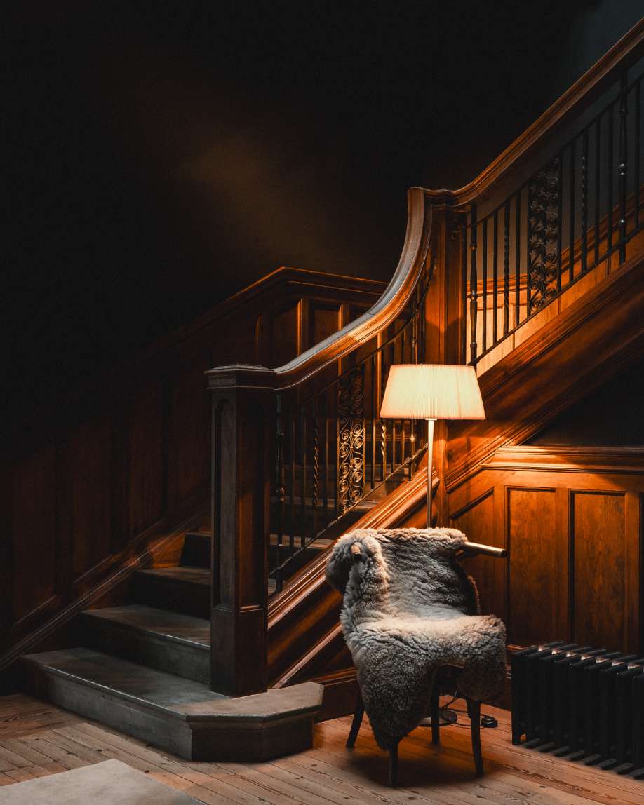 A sheepskin-draped armchair stands beside a lit lamp at the foot of a dark wood staircase with panelled walls.