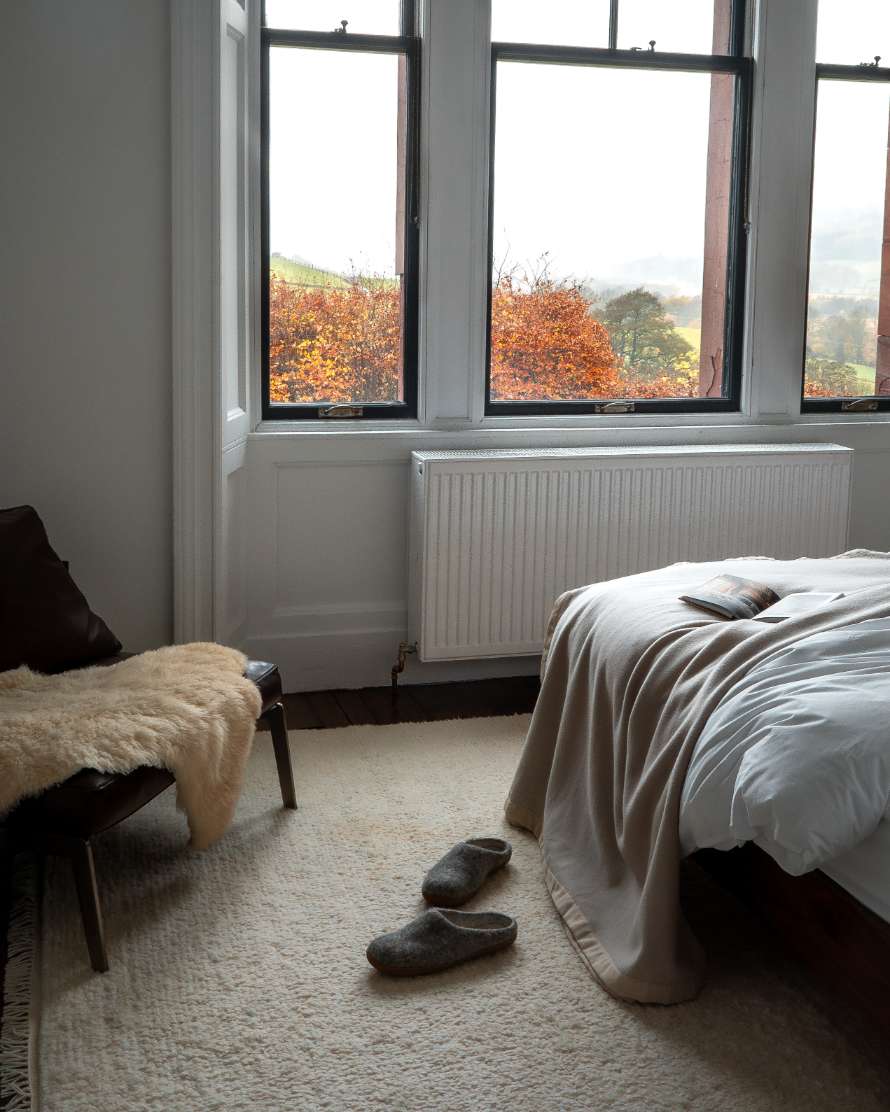 A bed dressed with a cream throw stands below a three-pane window looking out to autumn trees, grey slippers on the rug and a leather bench with a sheepskin nearby.