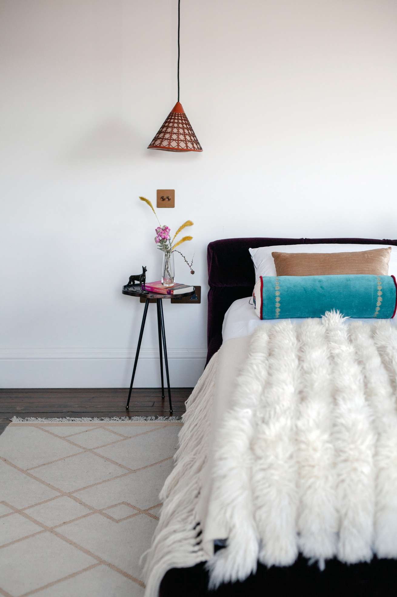 A bed with a dark velvet headboard is dressed with a teal bolster cushion, a tan cushion and a shaggy fur throw, a pendant lamp hanging above a side table of flowers and books.