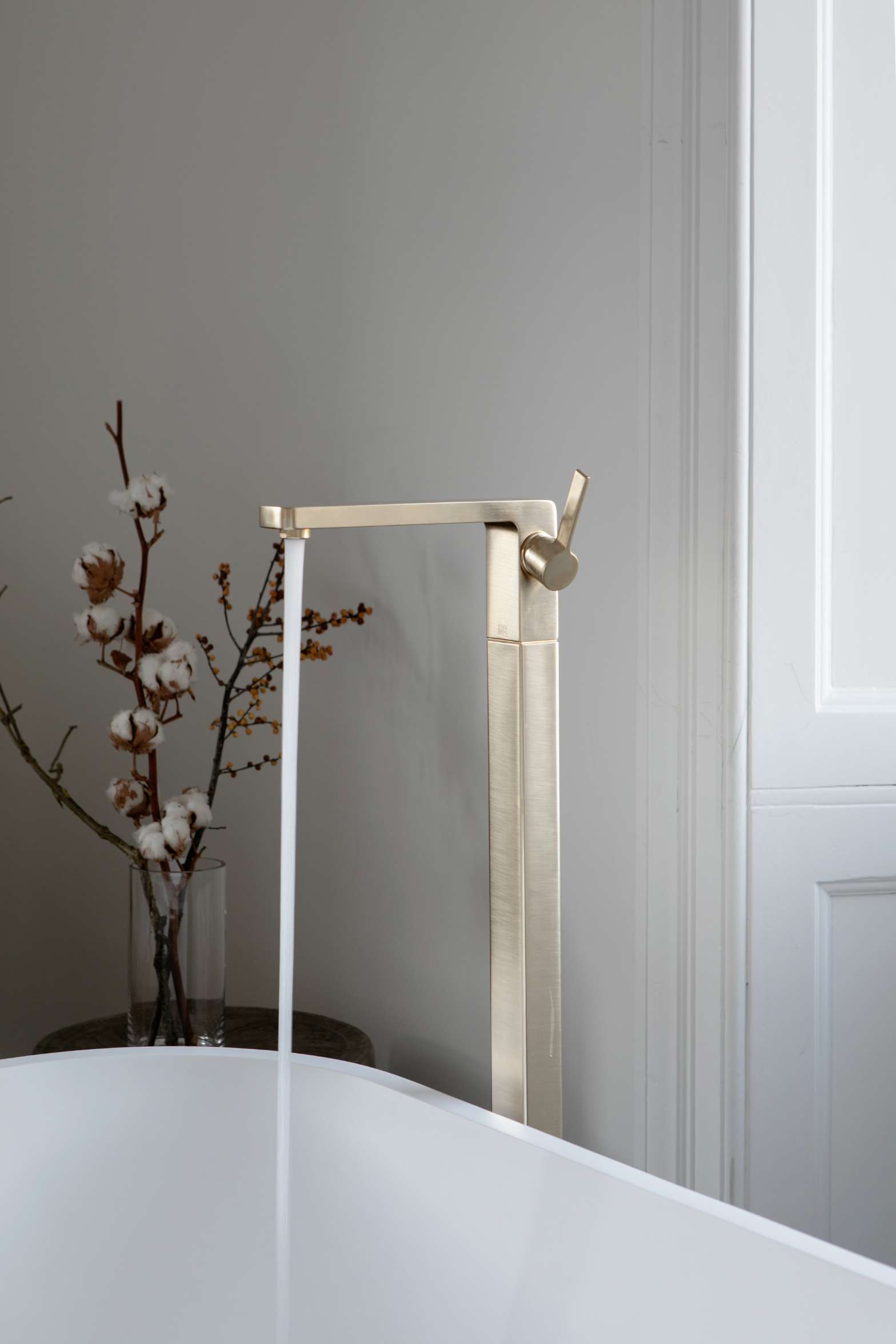 Water pours from a gold-toned tap into a freestanding white bath, a vase of cotton stems on a small table beside it.