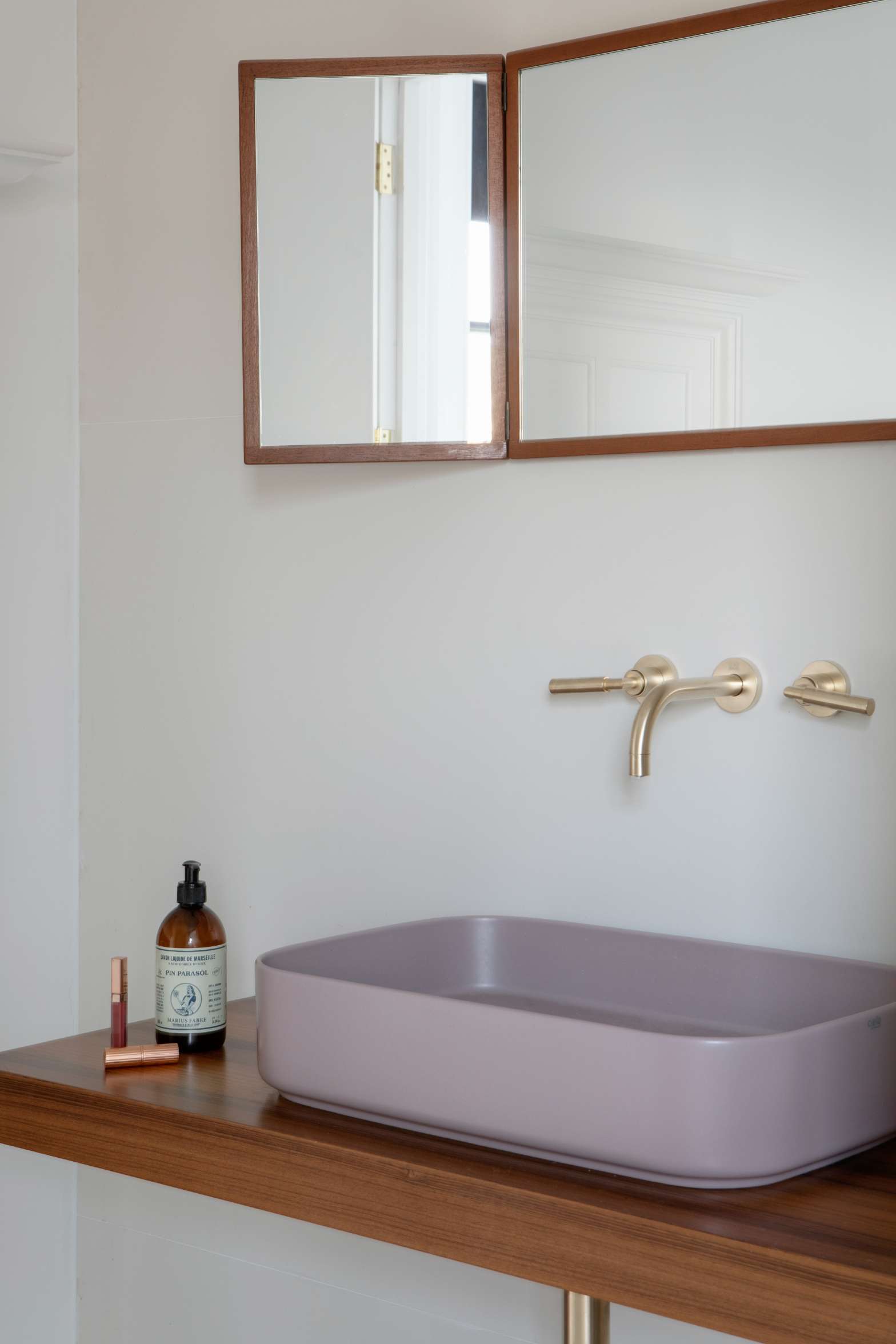 A mauve oval basin sits below gold-toned wall taps and a mirrored cabinet, a bottle and lipsticks set on the wooden counter beside it.