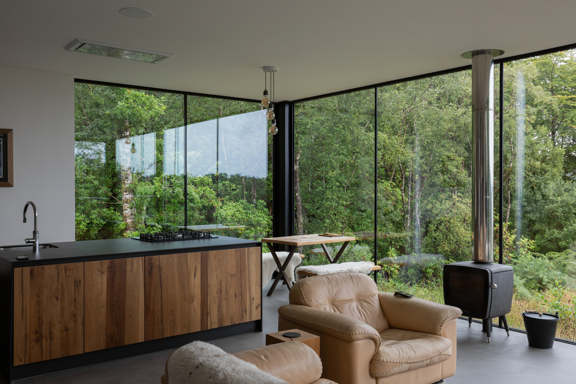 A leather armchair with a sheepskin throw sits beside a wood burning stove, a dark kitchen and dining table visible before floor-to-ceiling windows onto woodland.