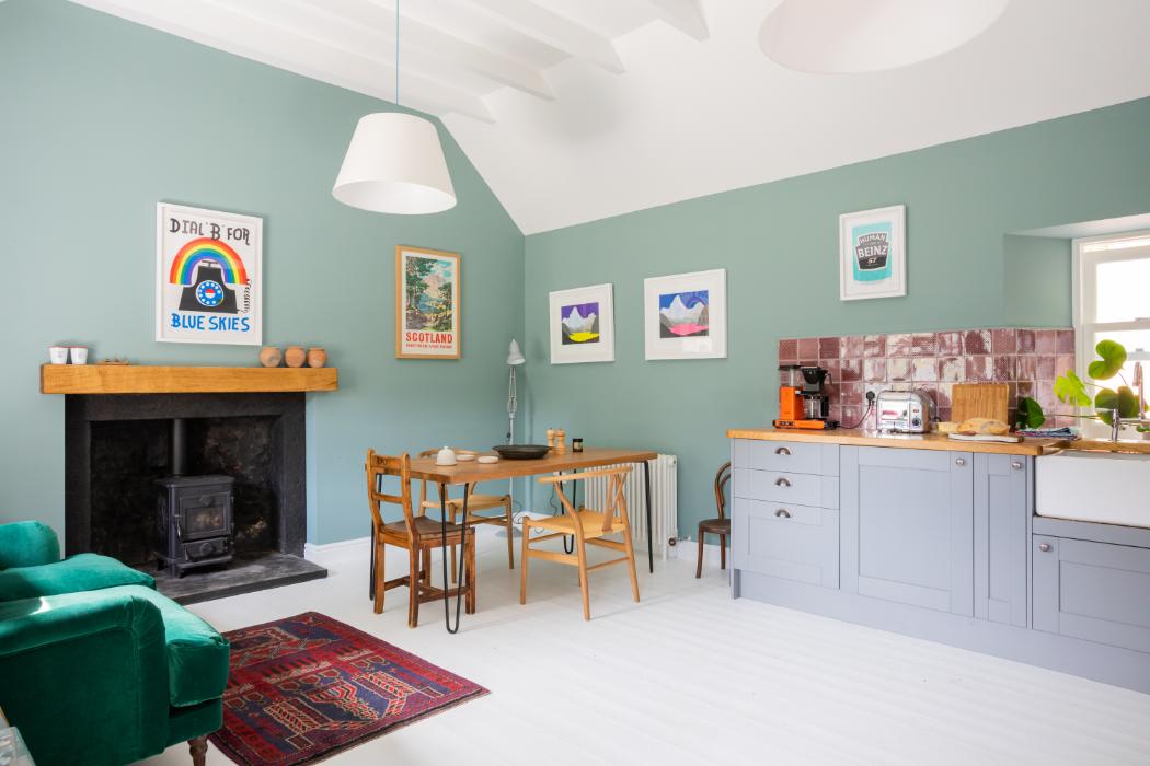 A green sitting and dining room holds a wood burning stove beneath a mantel of framed travel prints, a wooden table and grey kitchen units to the right.