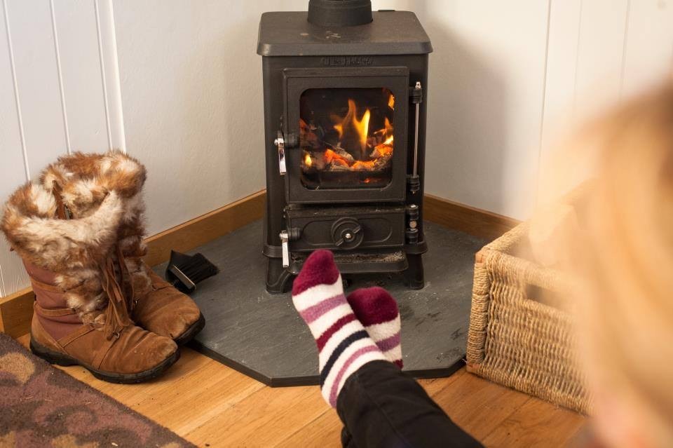 A pair of striped socks warm by a lit wood burning stove, fur-lined boots and a woven basket resting on the hearth beside it.