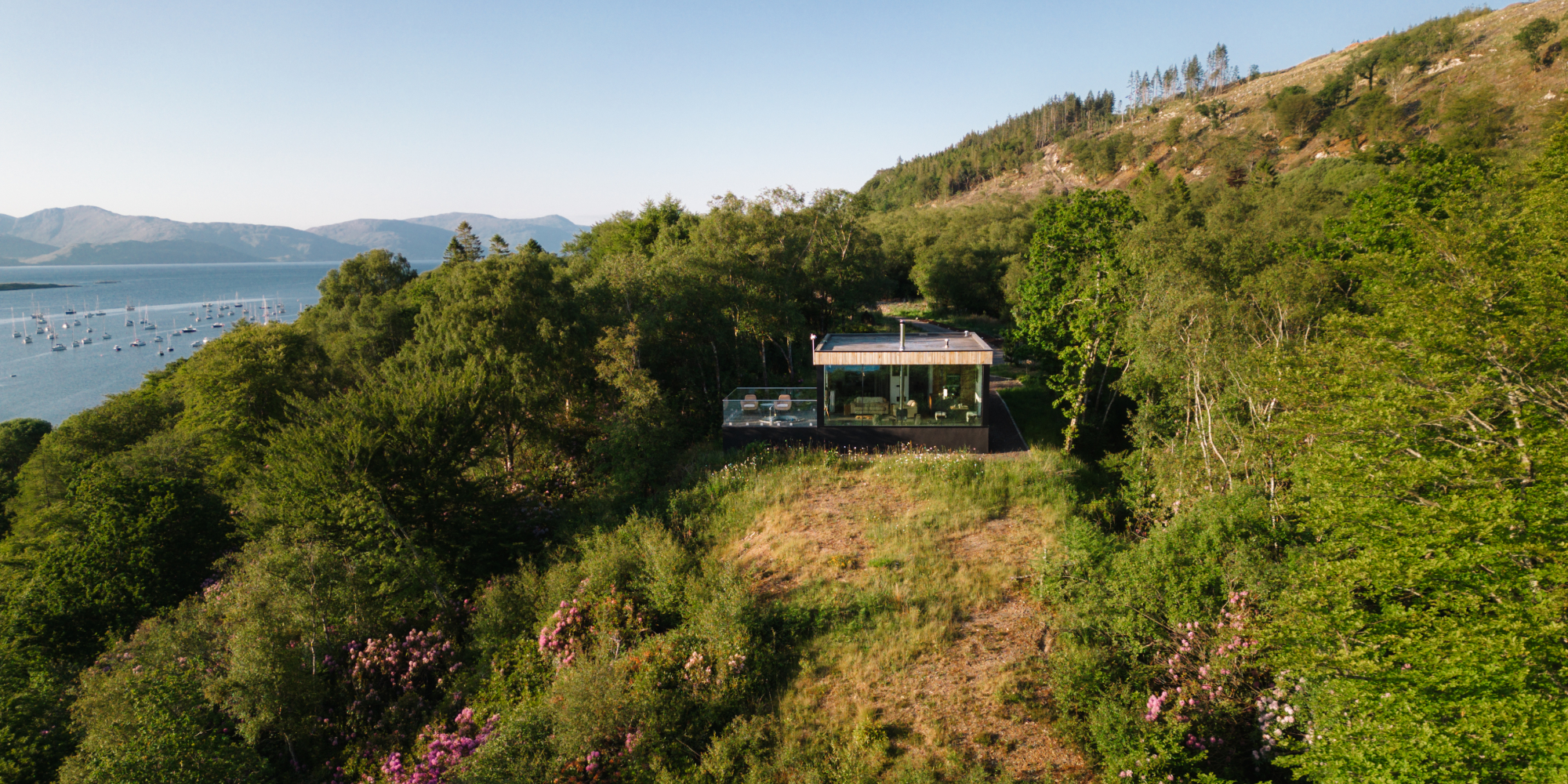 An aerial view shows a cedar-clad house nestled among trees and rhododendrons on a hillside above a loch.