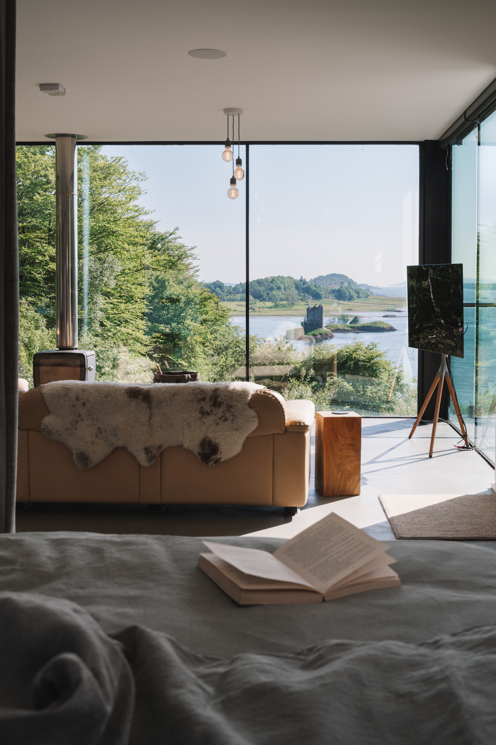 An open book lies on a bed before a leather sofa with a sheepskin throw, floor-to-ceiling windows framing a loch and a ruined tower on an islet.