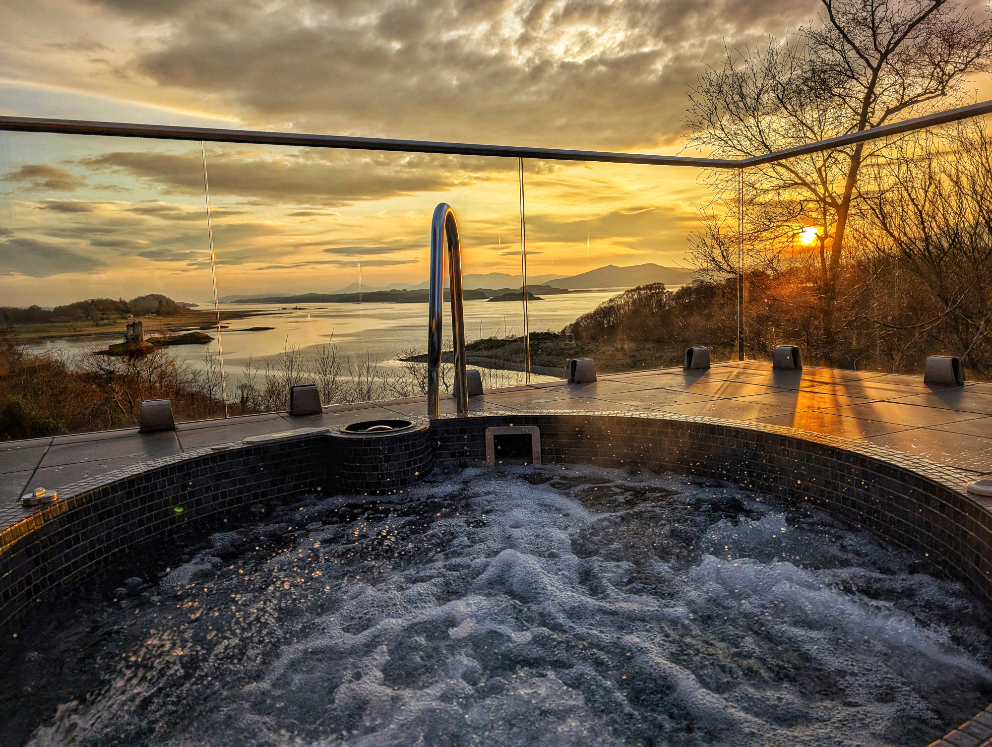 A hot tub bubbles on a stone terrace at sunset, an island and hills visible across the water through a glass balustrade.