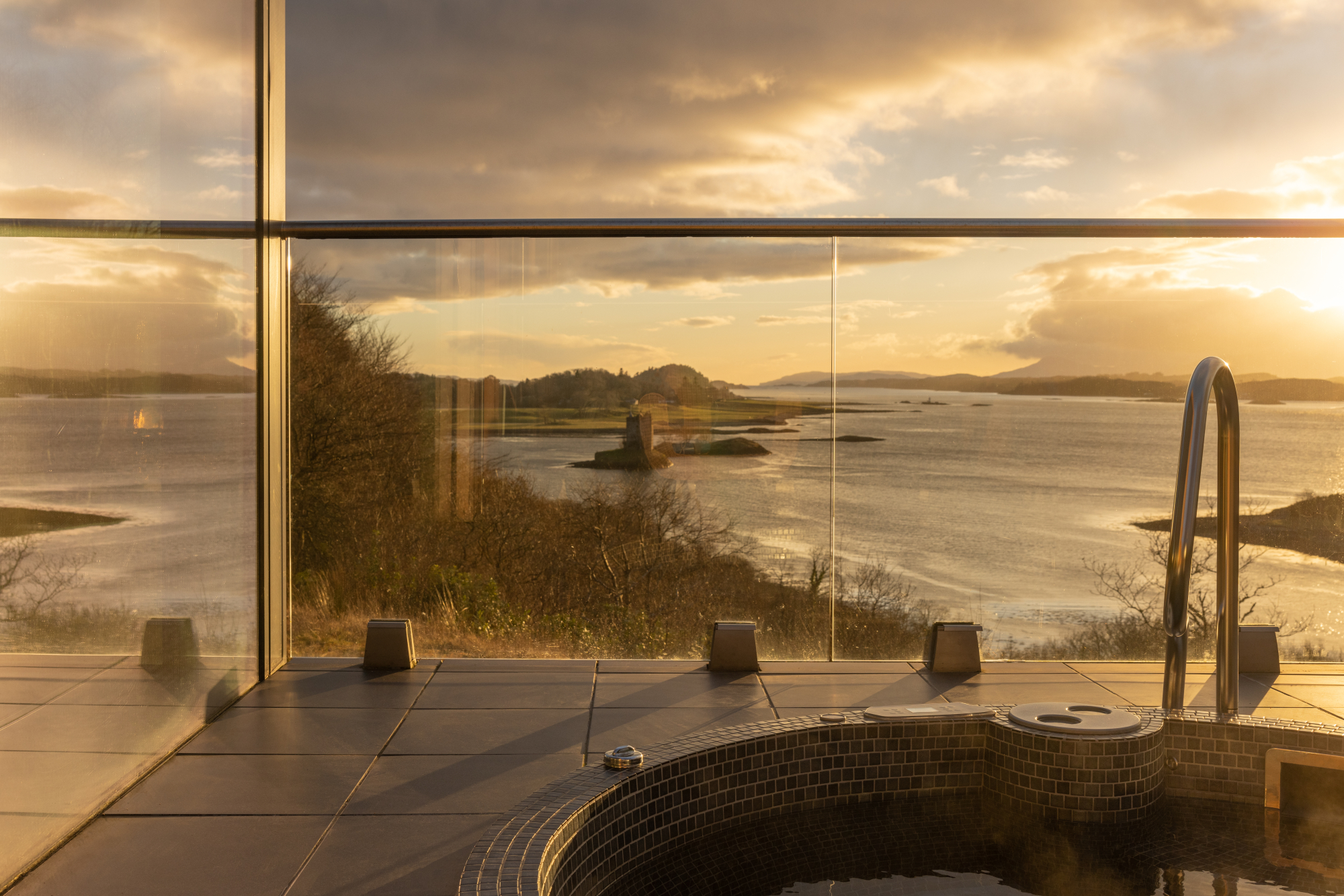 A sunken hot tub with a chrome ladder looks out over a loch scattered with islands and a ruined tower at sunset.