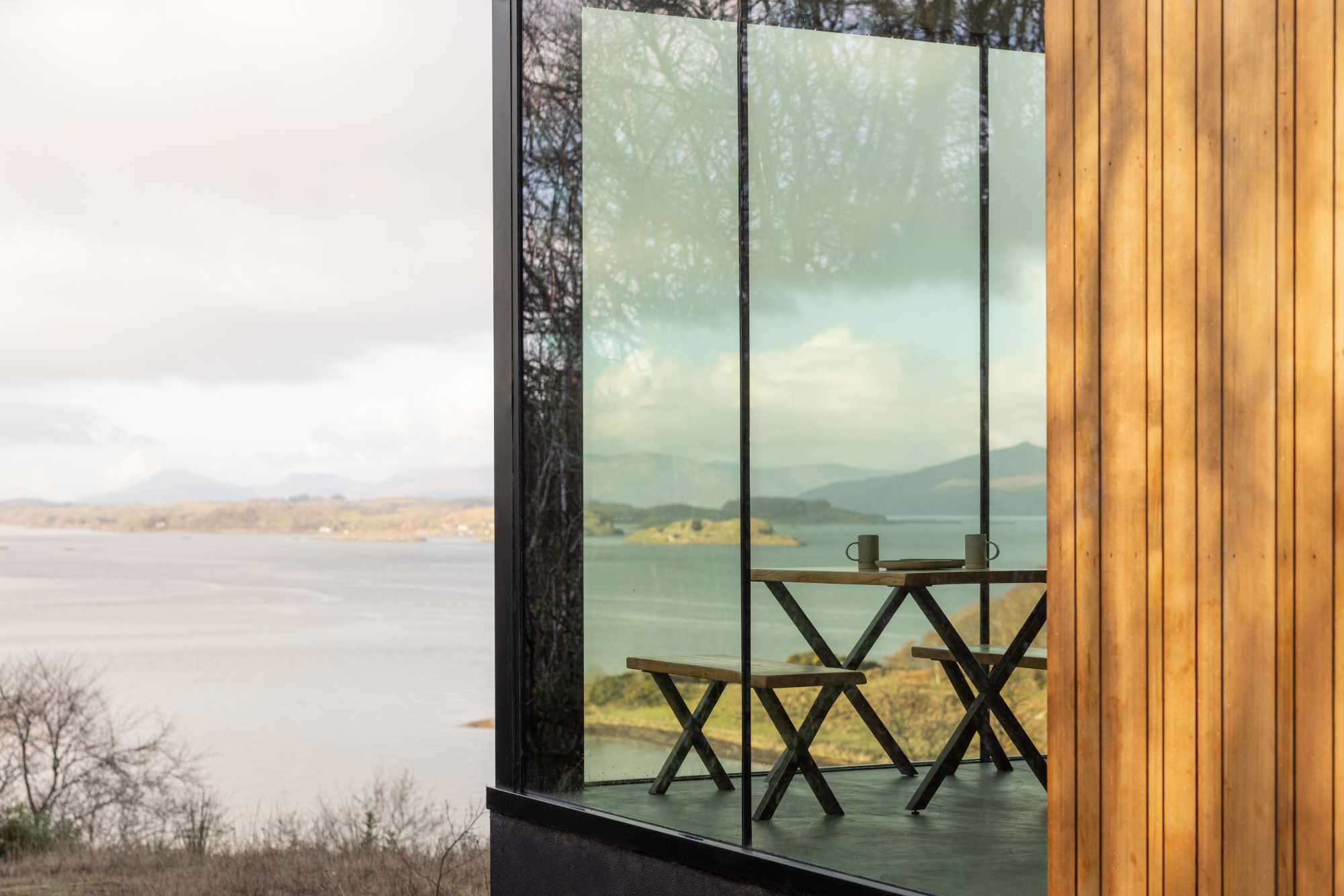 A wooden picnic table is reflected in a glazed corner window, a loch and hills visible beyond the cedar cladding.