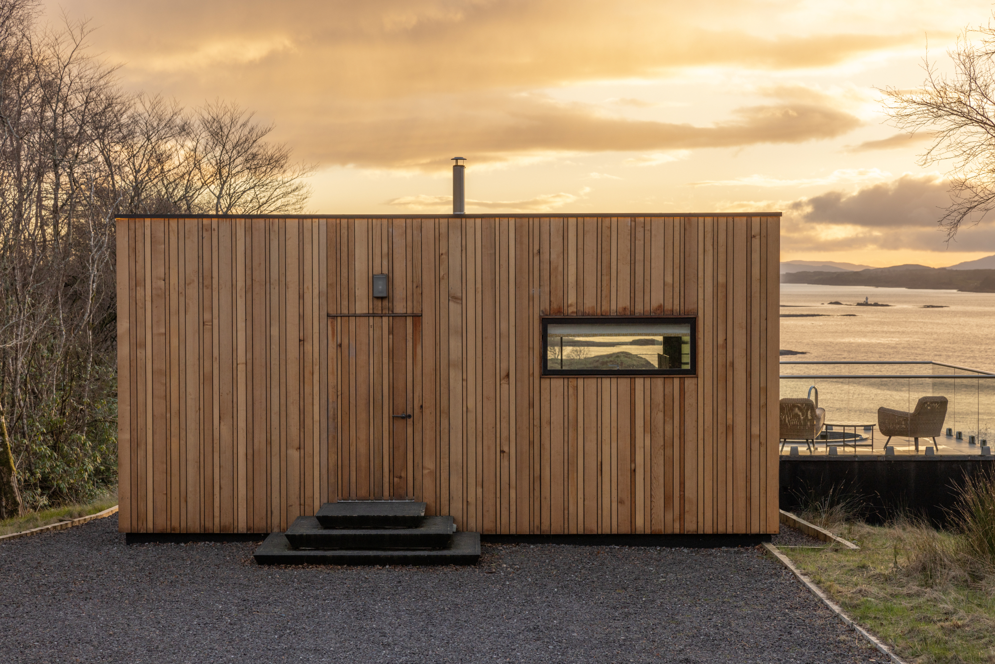 A cedar-clad cabin with a stove flue catches the sunset glow, steps leading up to its door and a window framing hills and water.