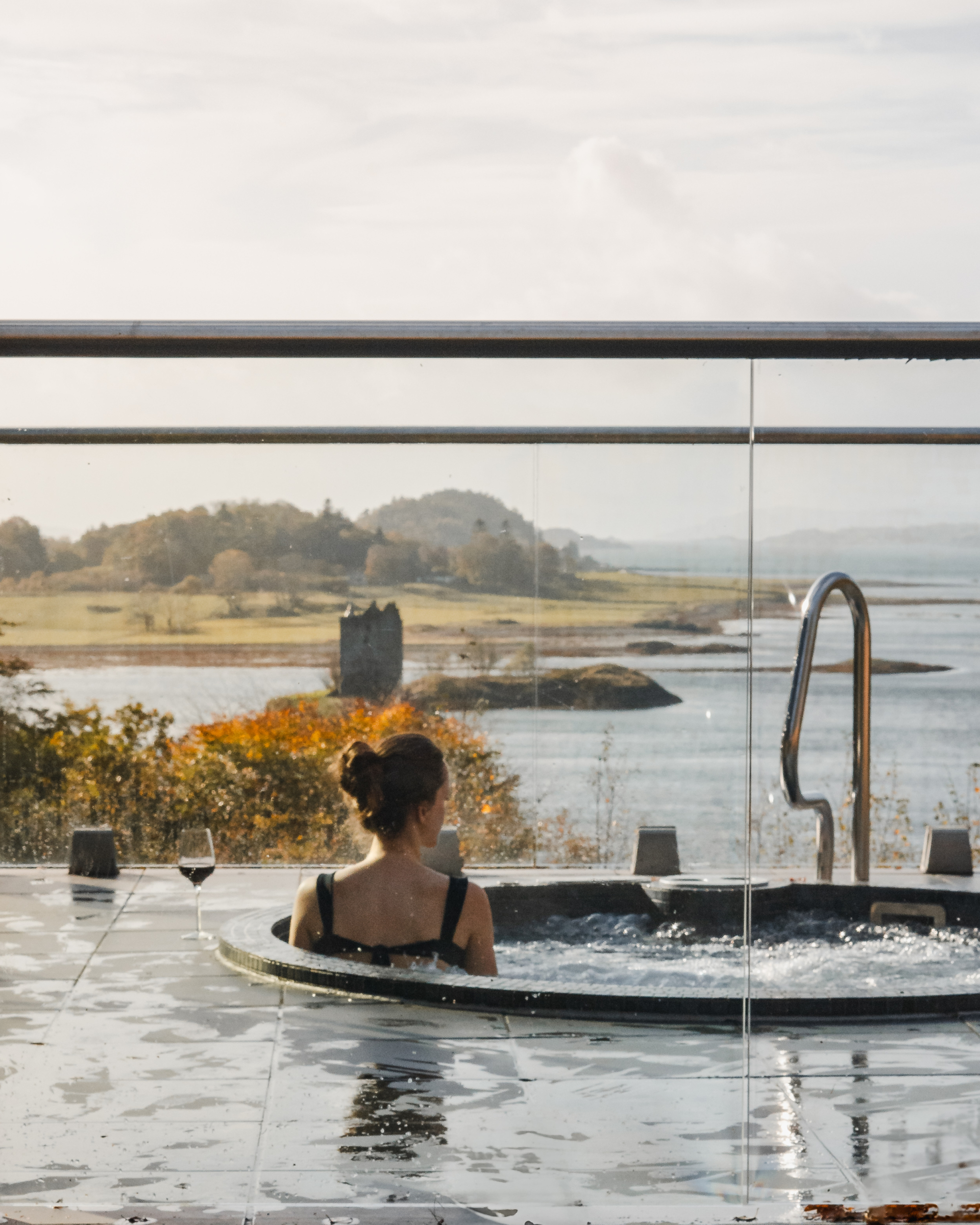 A woman relaxes in a hot tub with a glass of wine, a ruined tower on an islet visible across the water beyond the glass balustrade.