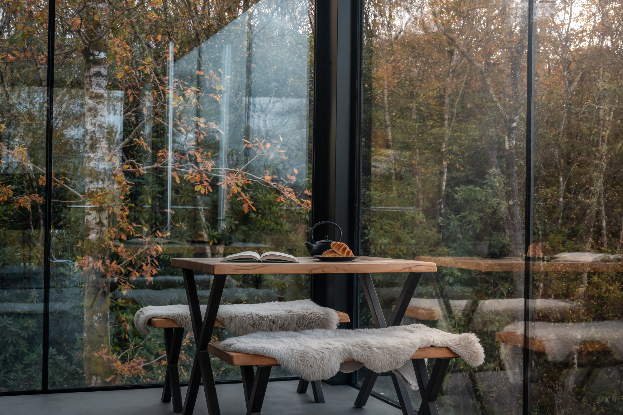 A round wooden table with sheepskin-topped stools stands before floor-to-ceiling glazing, a teapot and open book set on top among autumn woodland.