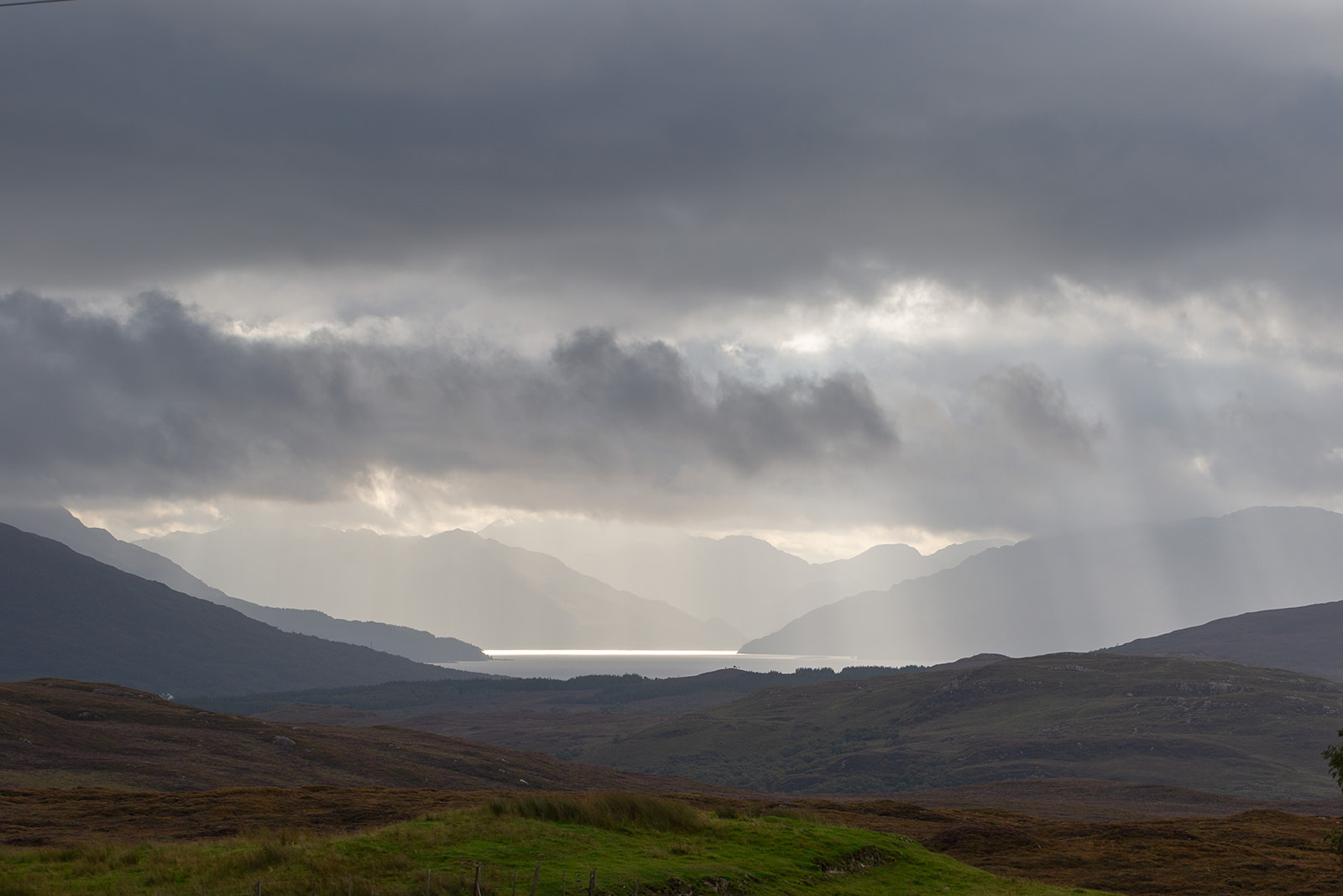 Sunbeams break through heavy cloud over a loch framed by hills and moorland.