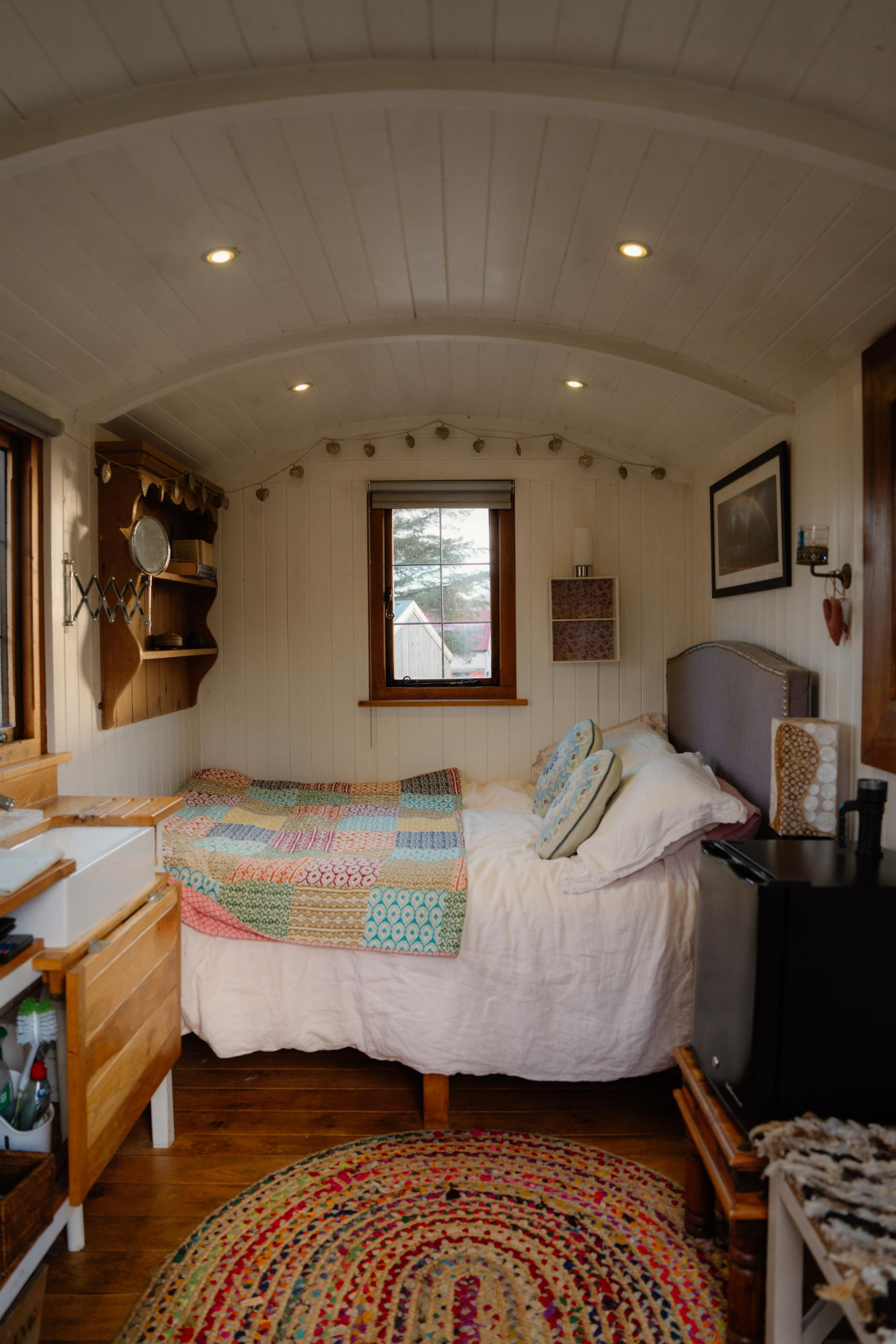 A bed with a patchwork quilt sits beneath a curved wooden ceiling strung with lights, a window looking onto a garden and a small dressing area to the left.