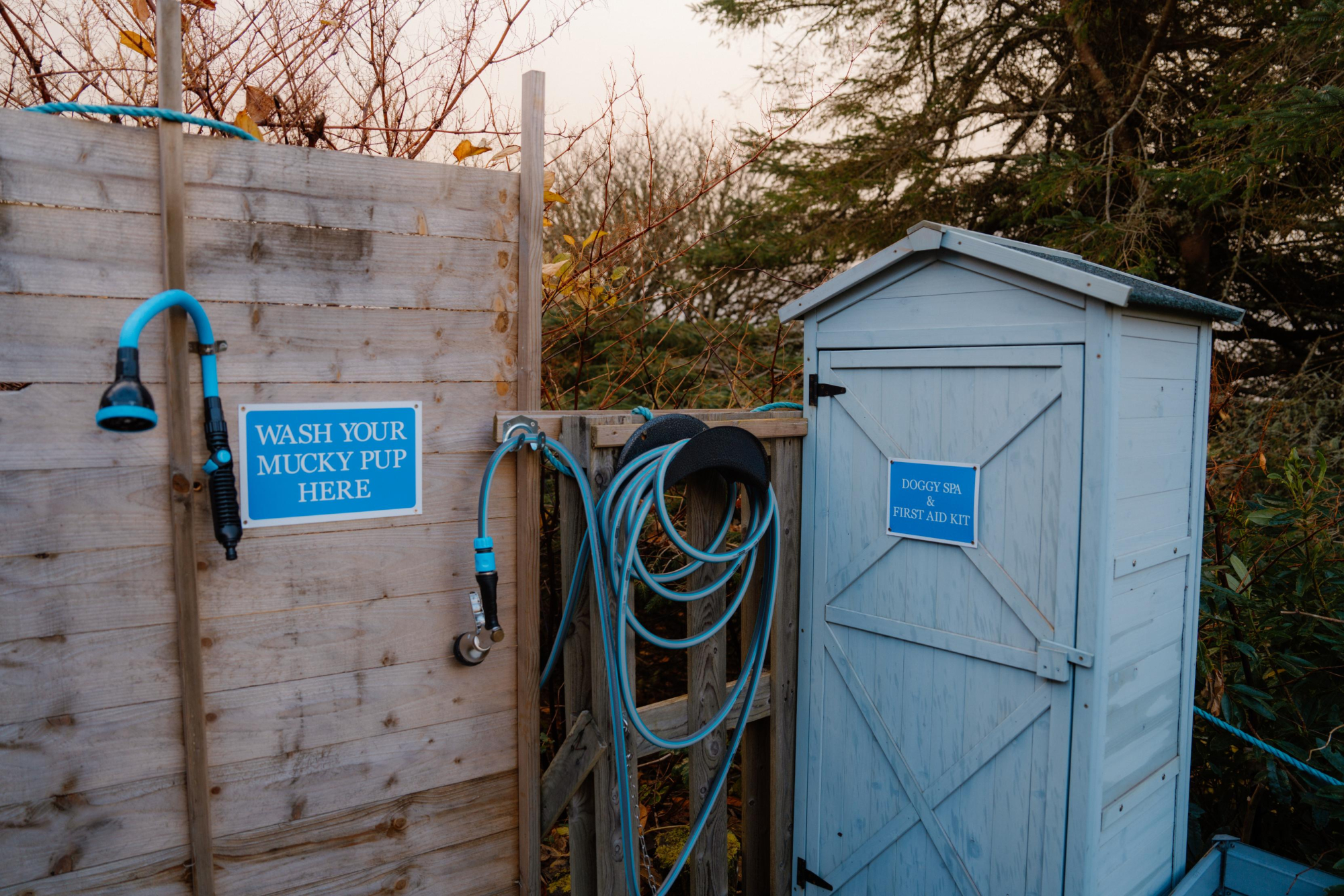 A sign on a timber fence reads "Wash Your Mucky Pup Here" beside a hose and shower head, a small outbuilding to the right.