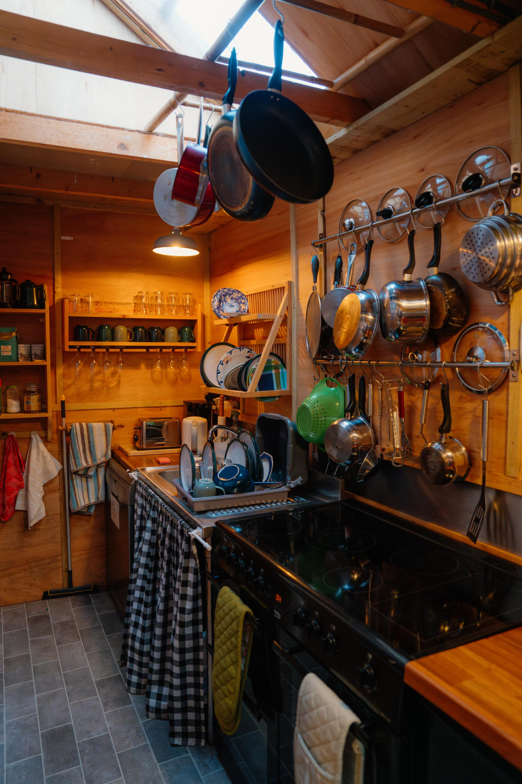 A galley kitchen under a skylight has pots and pans hanging above the stove, glasses and mugs on open shelving to the side.