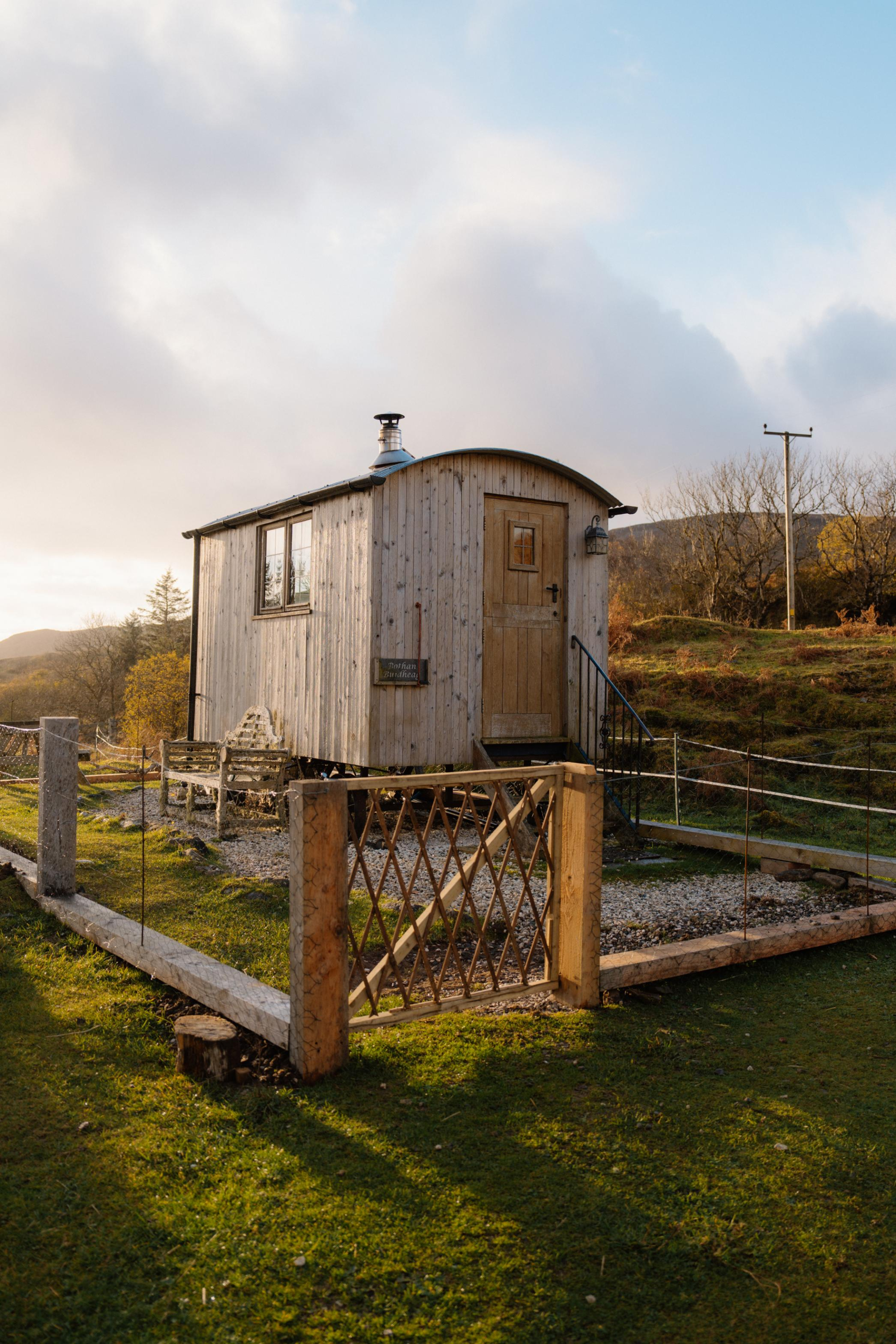 A timber shepherd's hut with a curved roof stands behind a diamond-lattice gate, steps leading up to its door and a bench alongside.