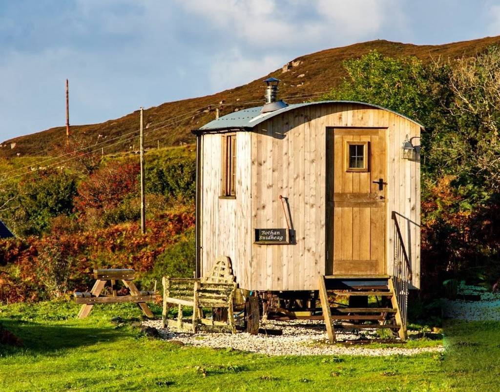 A timber shepherd's hut with a curved roof stands on a grassy slope, a bench and picnic table in front and hills rising behind.