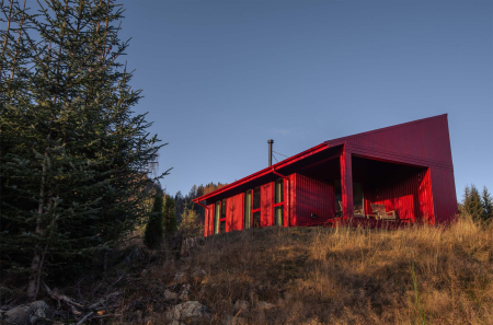 A red timber cabin with a steep pitched roof stands on a hillside among pines, a covered deck and chairs to one side.