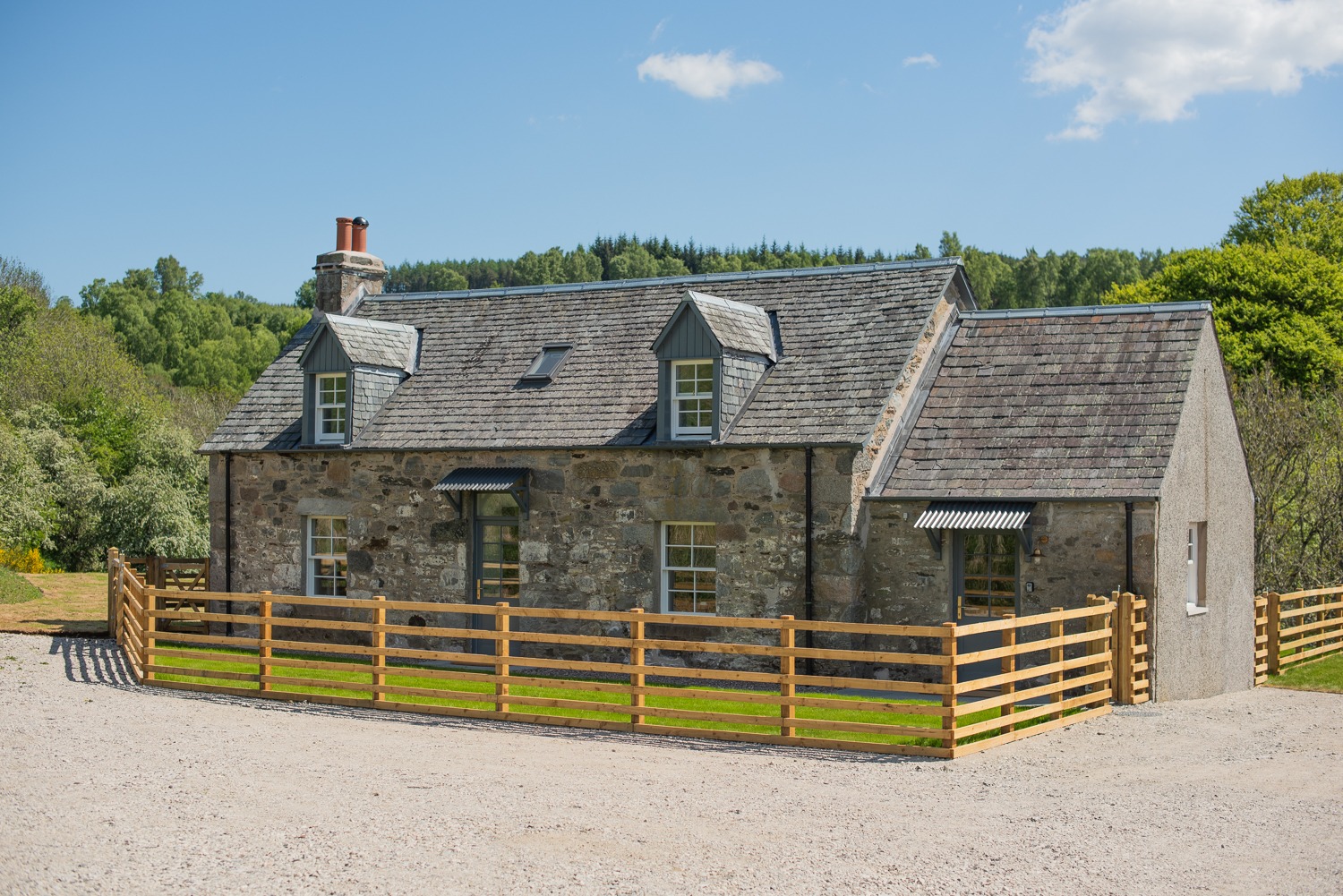 A stone farmhouse with dormer windows sits behind a timber post and rail fence, trees and hills visible beyond.