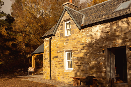 A stone cottage with a gabled dormer window catches evening light among autumn trees, a bench and picnic table on the gravel outside.