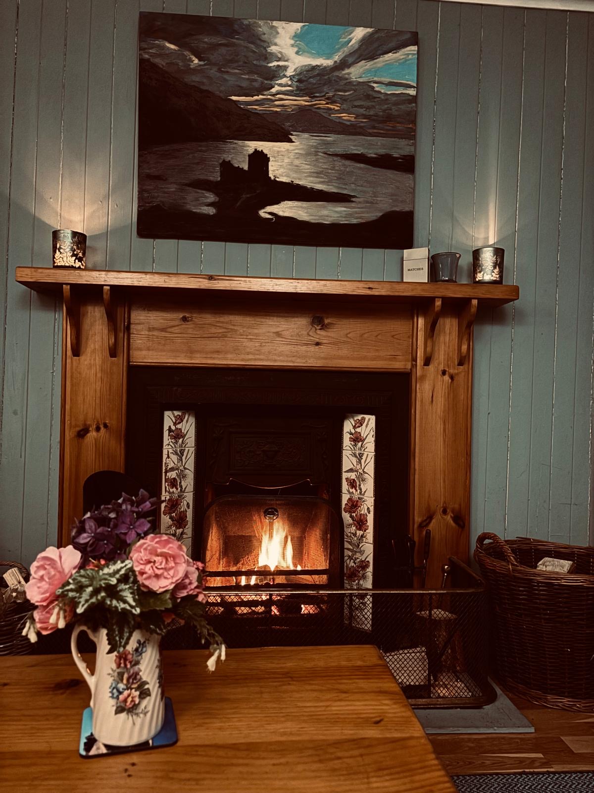 A painting of a castle on a loch hangs above a wooden mantel with a lit fire, a jug of pink and purple flowers on the table in front.