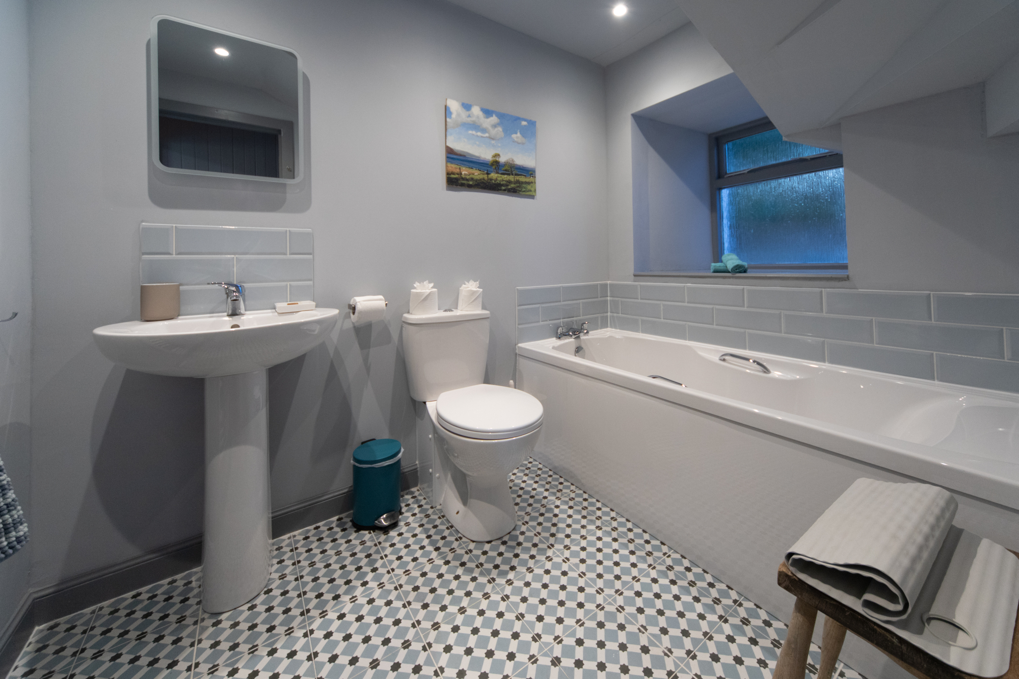 A pedestal basin and toilet stand beside a bath below a window, a patterned tiled floor and framed landscape print completing the bathroom.