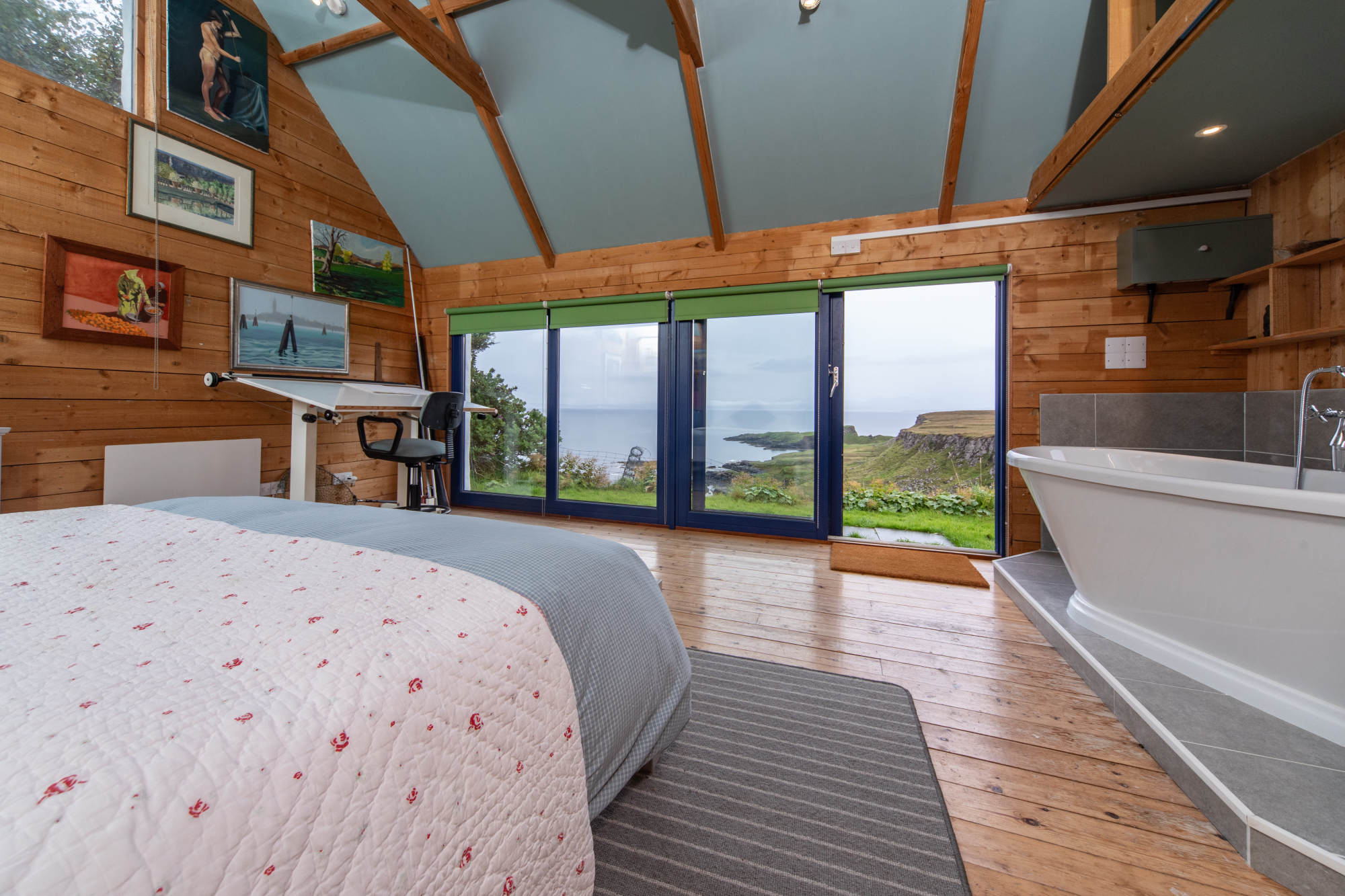 A bed faces glazed doors opening onto a sea view, a desk and chair to the left and a freestanding bath to the right under a vaulted timber ceiling.