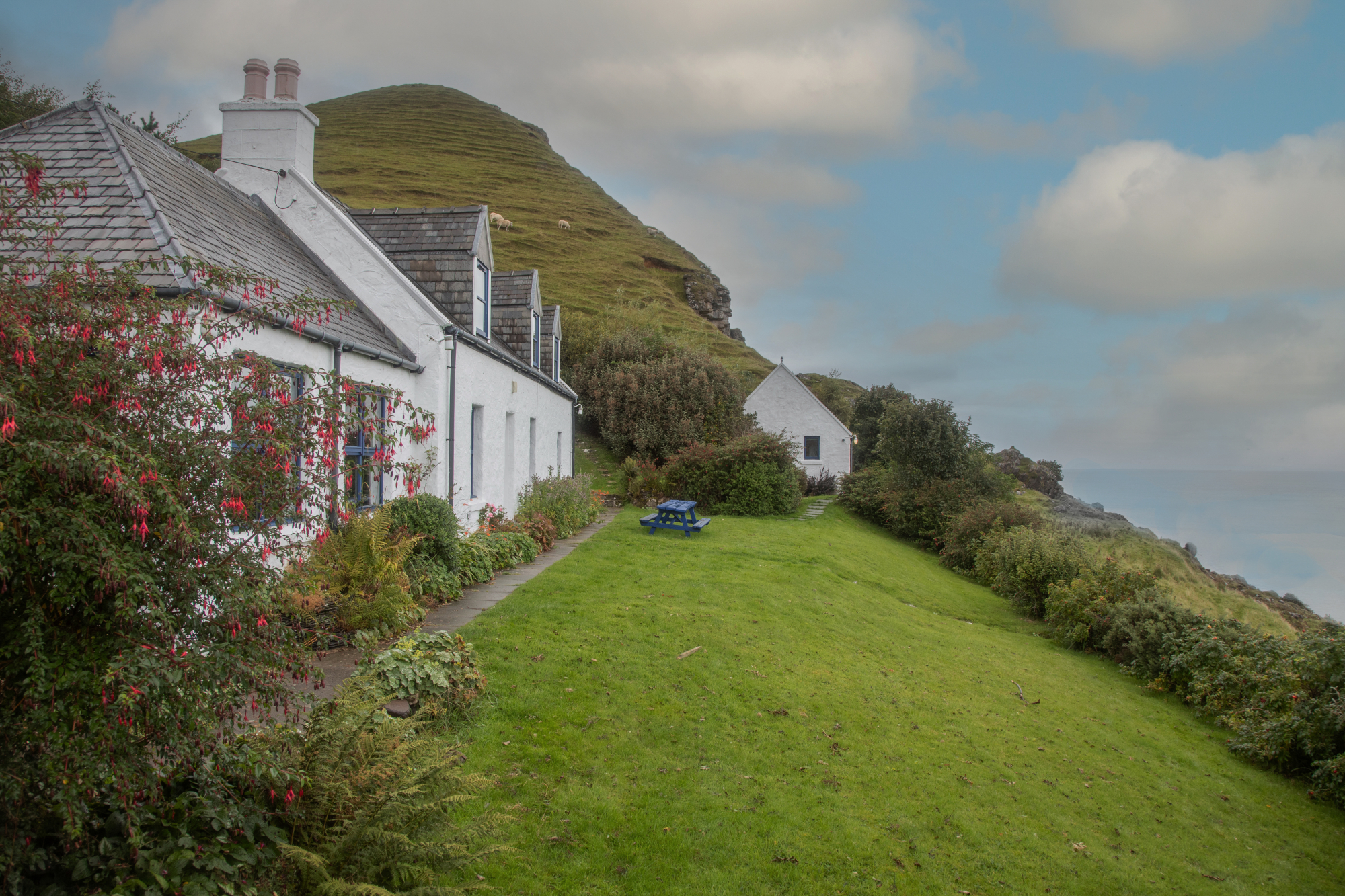 Fuchsia flowers climb the white walls of cottages beneath a grassy hill, a picnic table set on the lawn beyond.