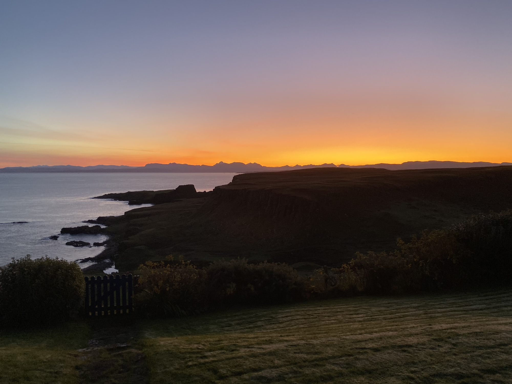 A wooden gate stands silhouetted against an orange sunset over a headland and calm sea.