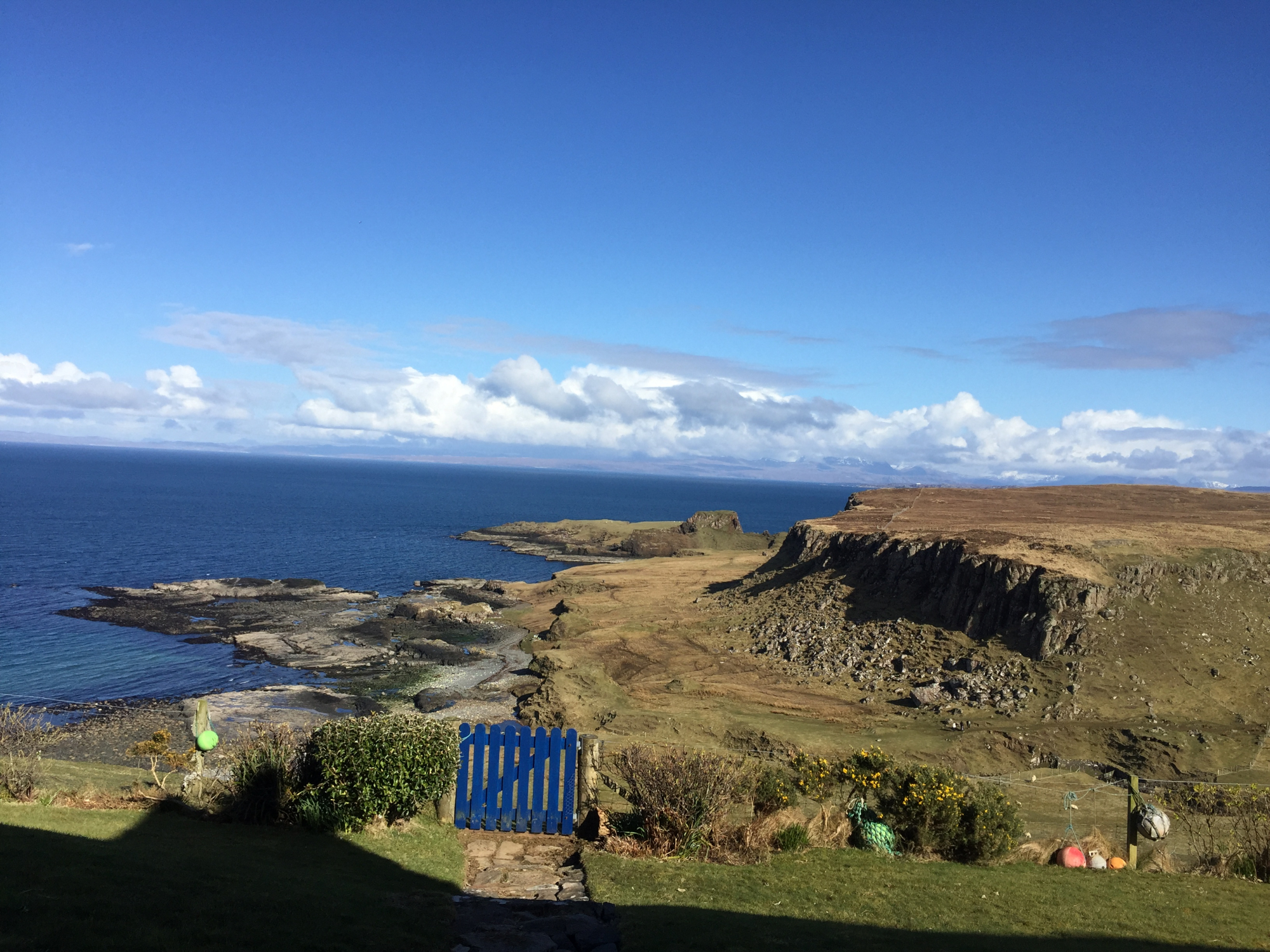 A weathered blue gate looks out over a rocky coastline and calm sea under a clear sky.
