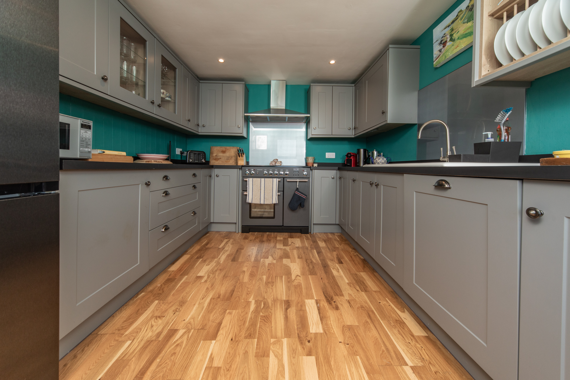 A grey kitchen with a teal splashback and range cooker, cabinetry running along both walls above a wooden floor.