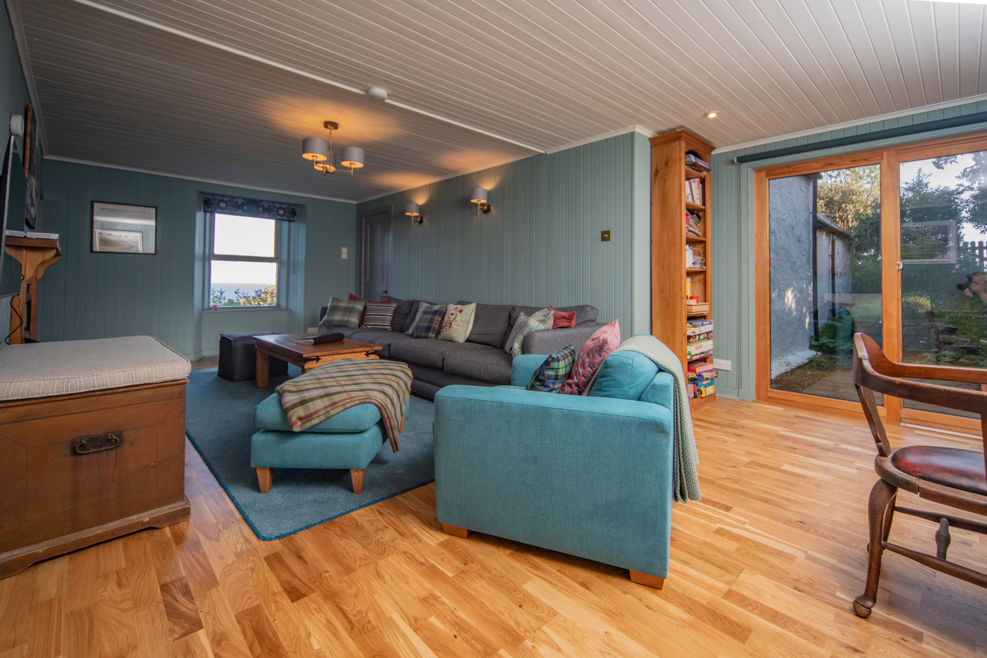 A teal corner sofa with cushions sits before a bookshelf and glazed doors onto a garden, a striped ottoman and antique chair in the foreground.