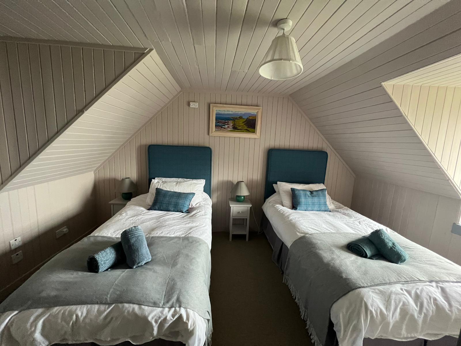 Twin beds with teal upholstered headboards sit either side of a bedside table, a framed coastal painting on the panelled wall between them.