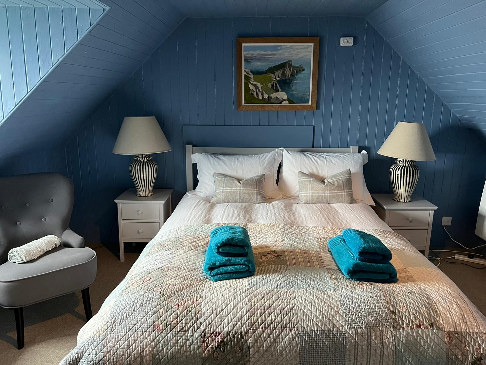 A bed with a patchwork quilt and rolled teal towels sits between two lamps on a blue panelled wall, a framed coastal painting above.