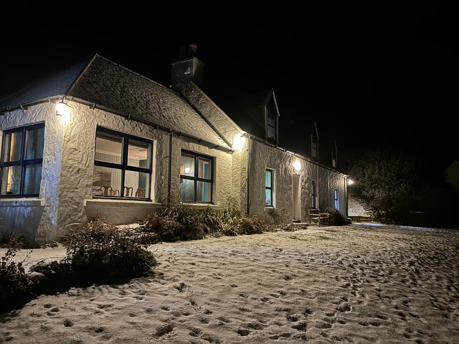 A white cottage glows with lit windows at night, snow dusting the ground and footprints crossing the drive.