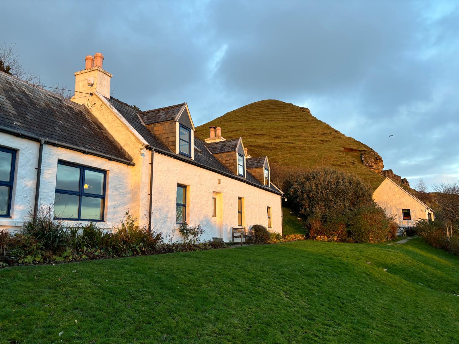 A row of white cottages catches the evening light below a grassy hill, a bench set on the lawn in front.
