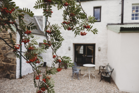 A rowan tree heavy with red berries hangs before a white harled house, a round table and chairs set on the gravel behind.