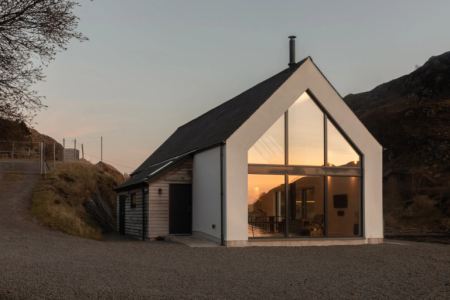 A gable-fronted white and stone house glows from within at dusk, its triangular glazed end lit against the darkening hillside.