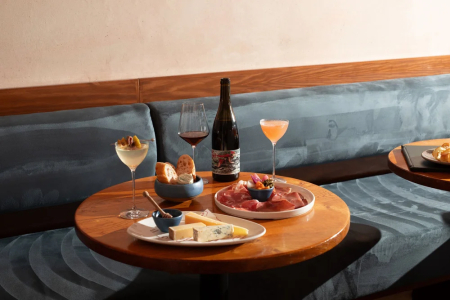 A round table set with wine, a cocktail, bread and charcuterie by a blue banquette.