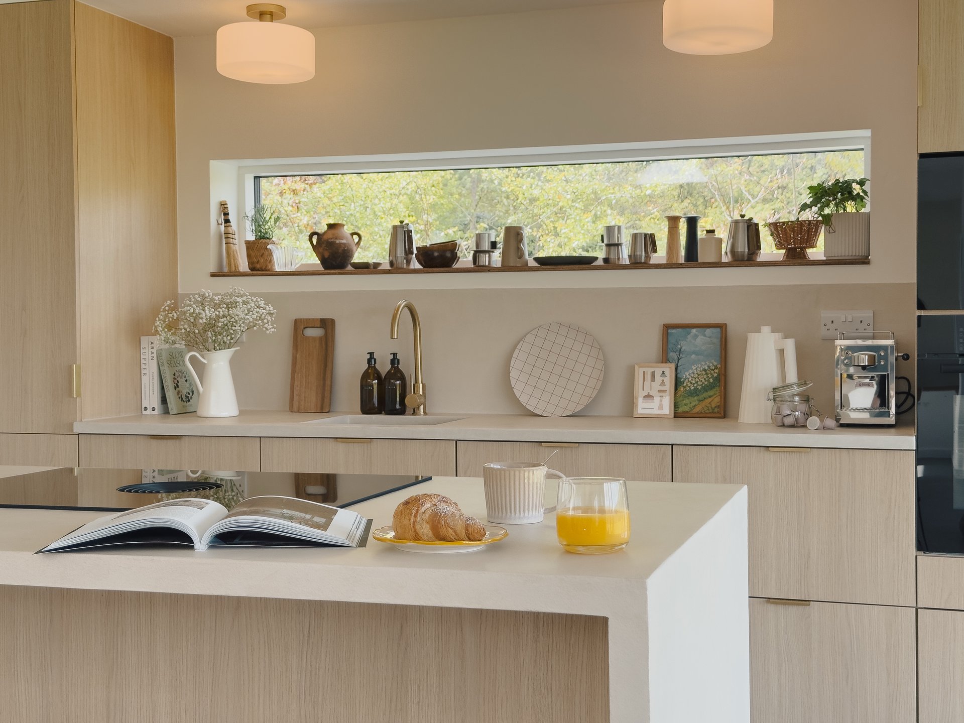 An open cookbook and a croissant sit on a kitchen island beside a glass of orange juice, a shelf of jugs and ornaments visible through the window above the counter.