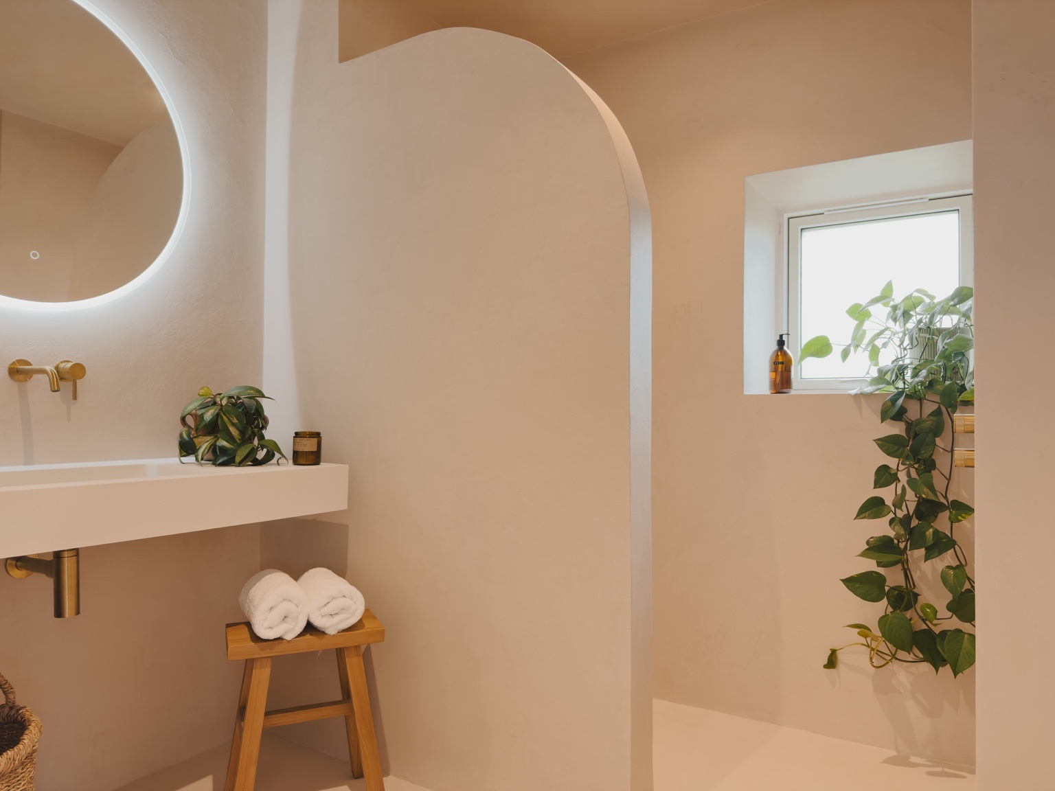 A wooden stool holds folded towels below a backlit round mirror, an arched doorway opening onto a trailing potted plant by a window.