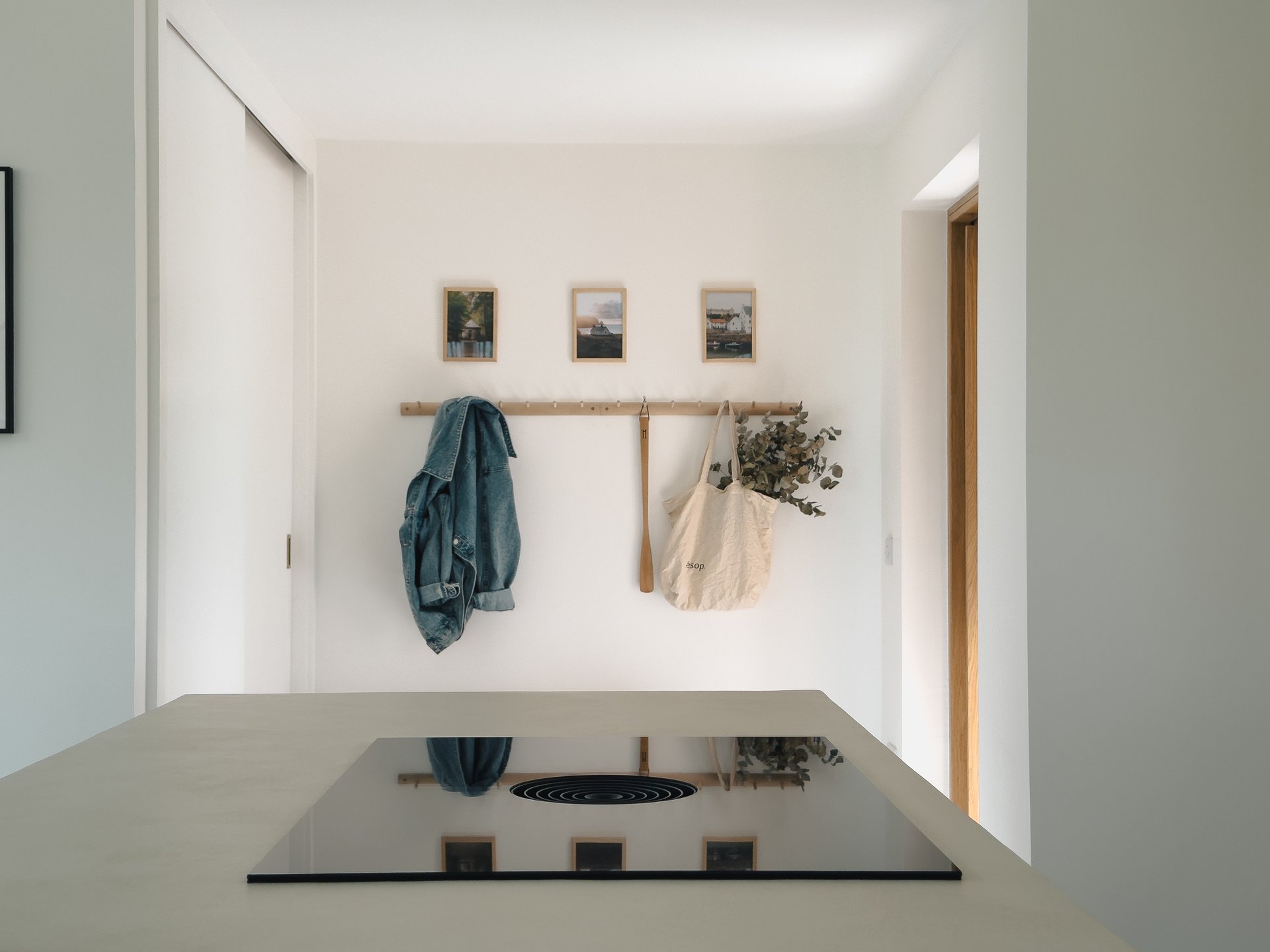 A denim jacket and tote bag hang from wooden pegs beneath three small framed photographs, a hallway leading to a door beyond.