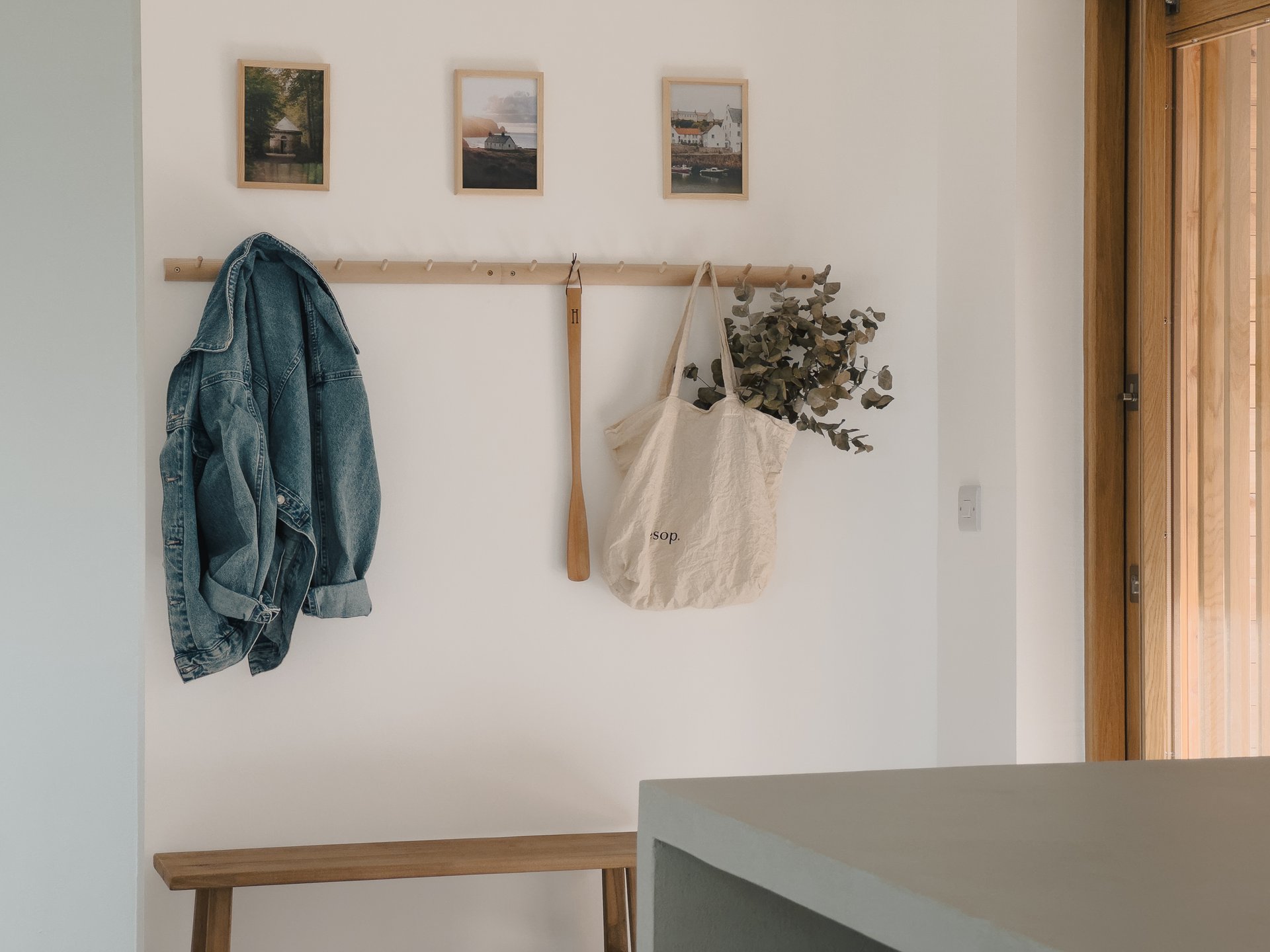 A denim jacket and canvas tote hang from wooden pegs below three small framed photographs, a bench and doorway to the side.