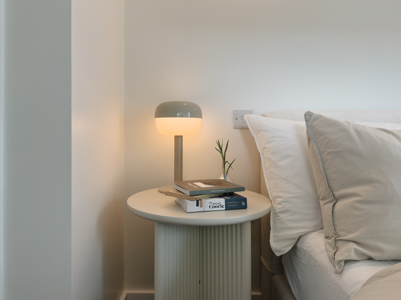 A round table lamp glows beside a stack of books and a sprig of foliage on a fluted side table, pillows on the bed to the right.