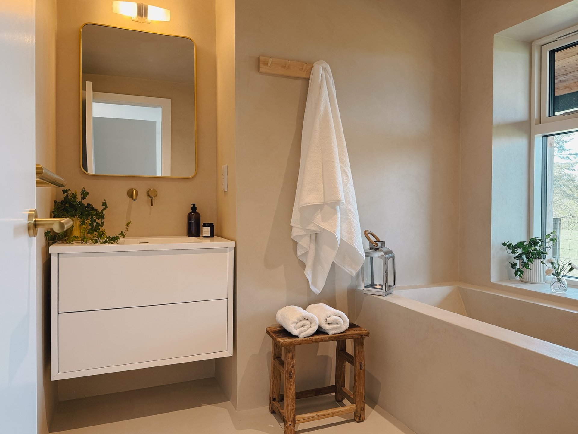 A floating vanity unit with a round mirror stands beside a towel rail hung with a robe, a wooden stool with folded towels in front of the bath.
